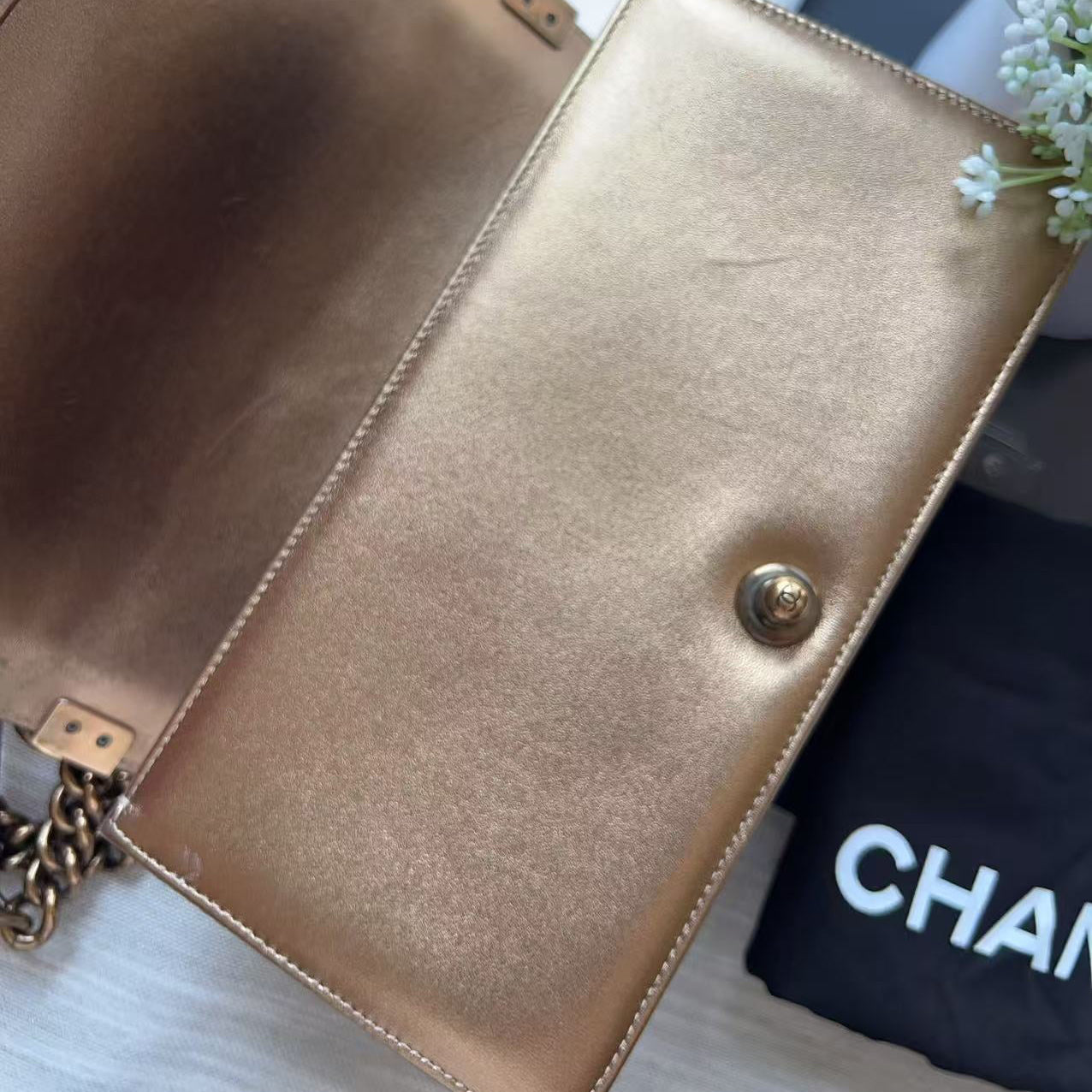 A metallic gold leather clutch bag shown open, with a gold-tone snap closure and chain strap, photographed on a light surface with a black Chanel dust bag and white flowers as props.