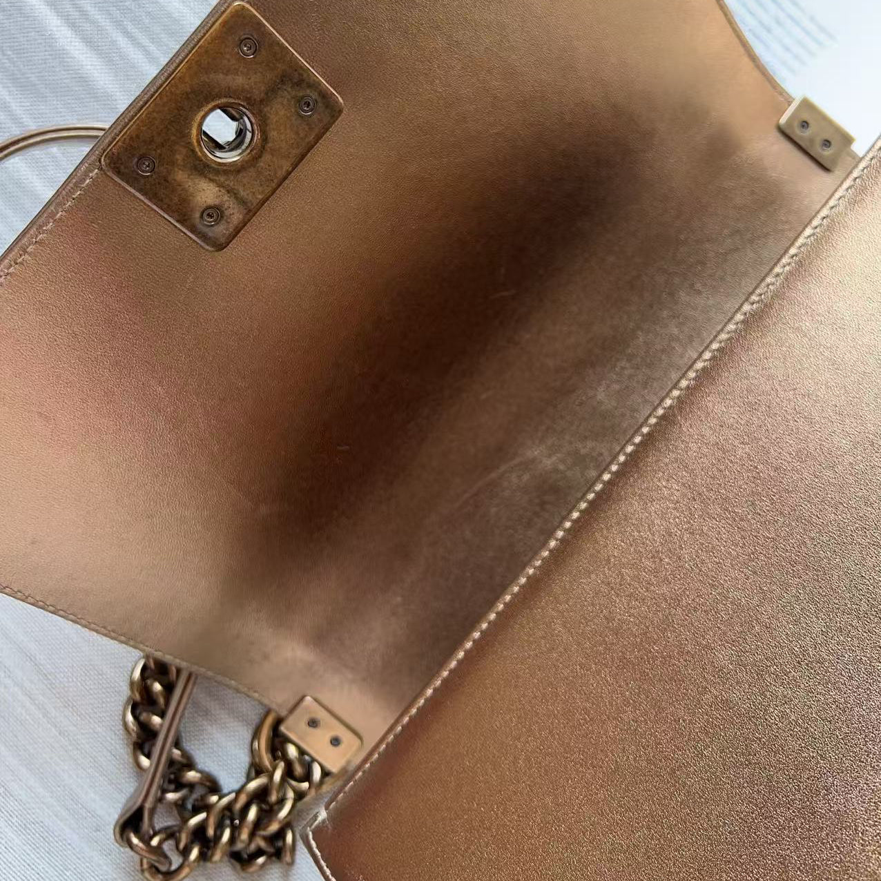 Close-up of a metallic rose gold leather clutch bag showing the interior flap, a central hardware lock, and a gold-tone chain strap on a light wood surface.