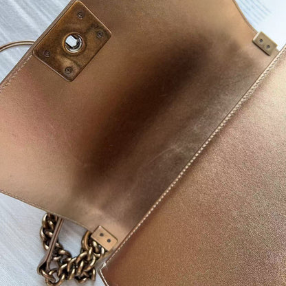Close-up of a metallic rose gold leather clutch bag showing the interior flap, a central hardware lock, and a gold-tone chain strap on a light wood surface.