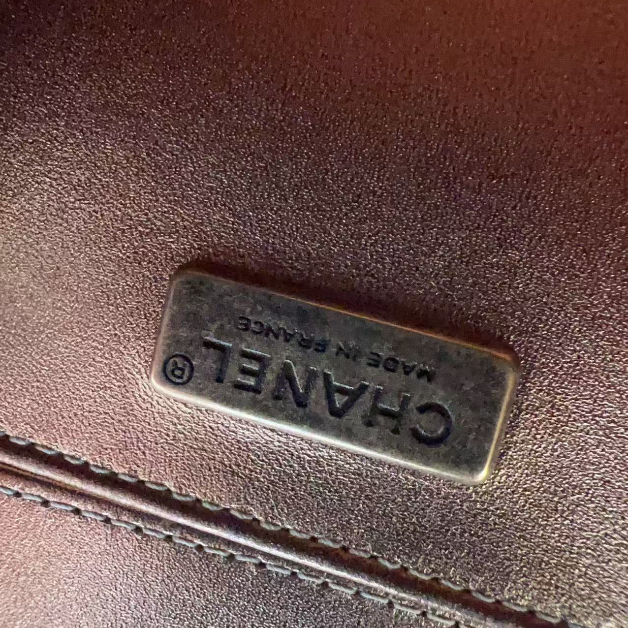 A close-up of a dark brown textured leather item featuring a rectangular metal plaque engraved with 'CHANEL MADE IN FRANCE'.