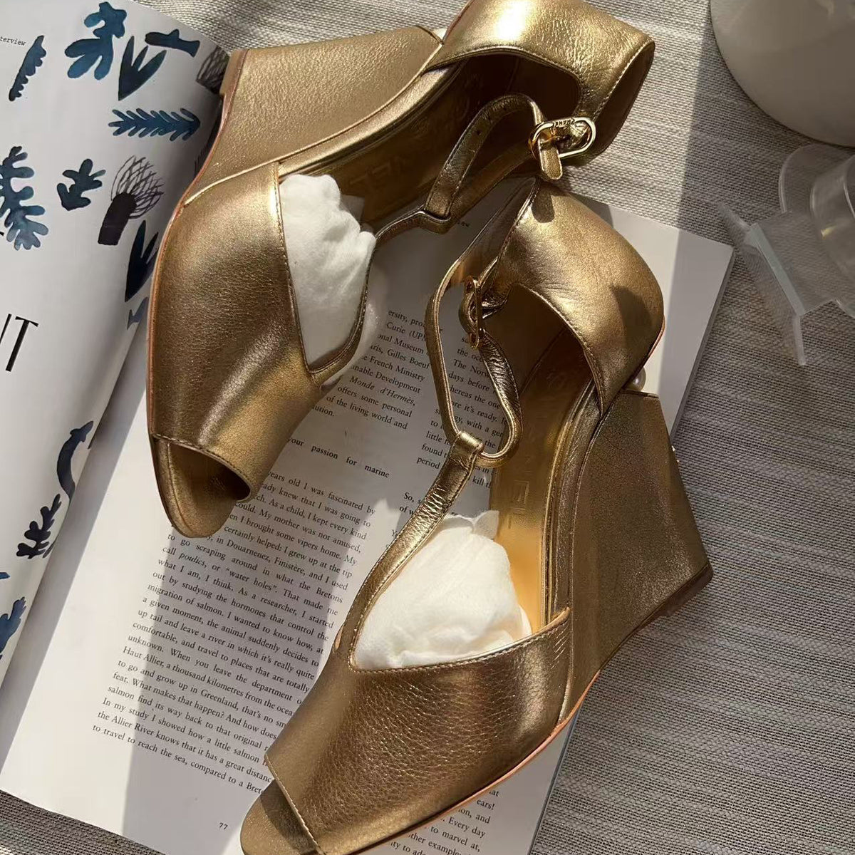 A pair of metallic gold wedge sandals with open toes and ankle straps, displayed on an open book with white tissue paper inserts. The background is a gray textured fabric with a white ceramic vase in the corner.