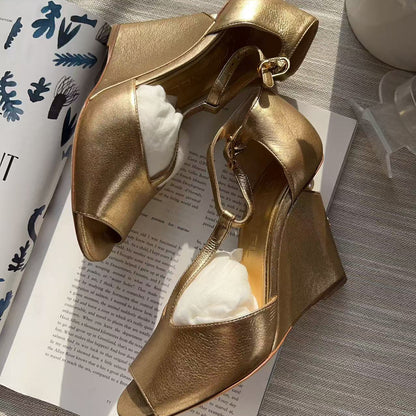 A pair of metallic gold wedge sandals with open toes and ankle straps, displayed on an open book with white tissue paper inserts. The background is a gray textured fabric with a white ceramic vase in the corner.