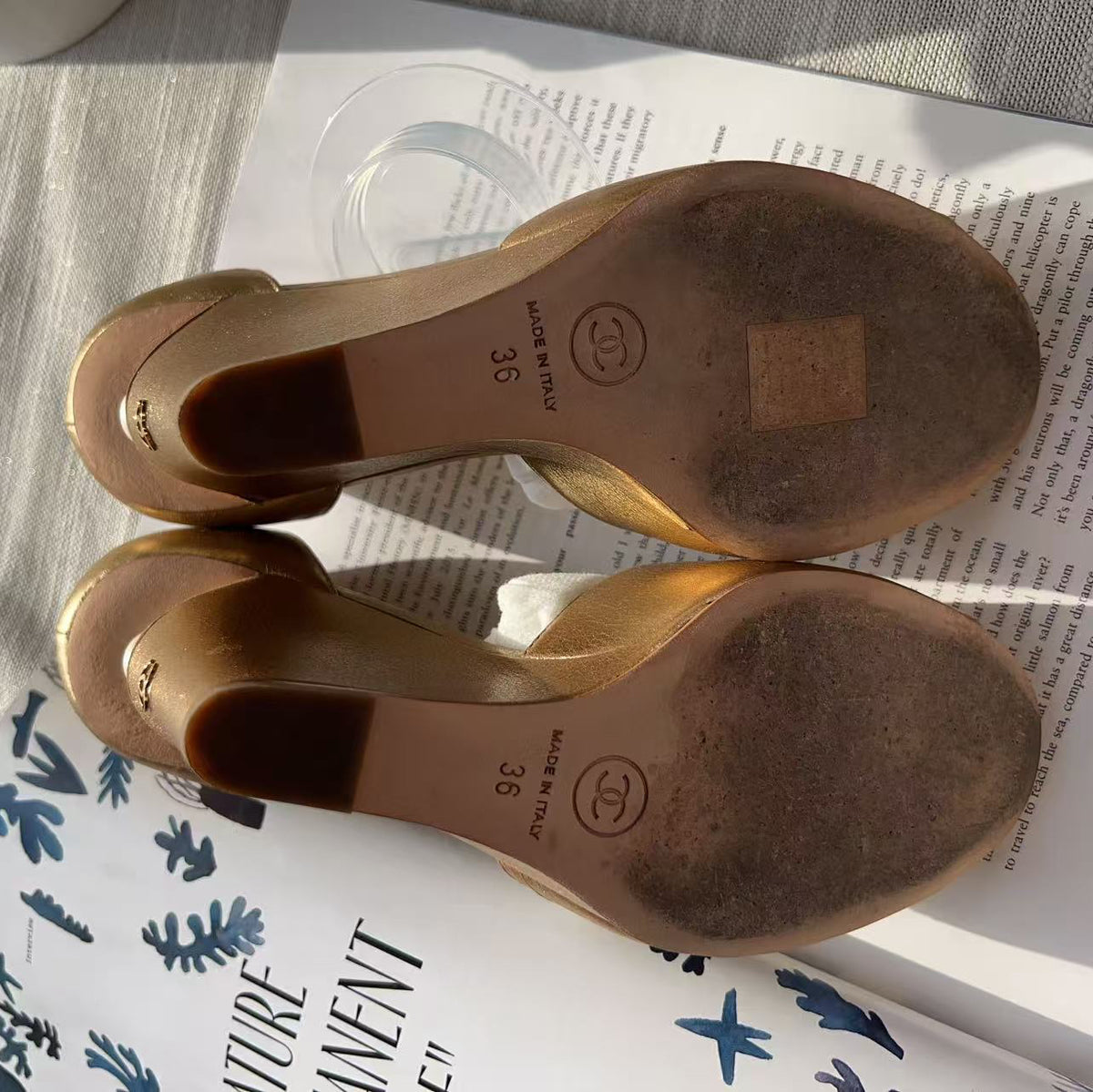 A pair of women's low-heeled sandals shown from the sole side, placed on top of printed papers over a light fabric surface. The soles are a warm tan-brown color with printed size markings and a circular logo.