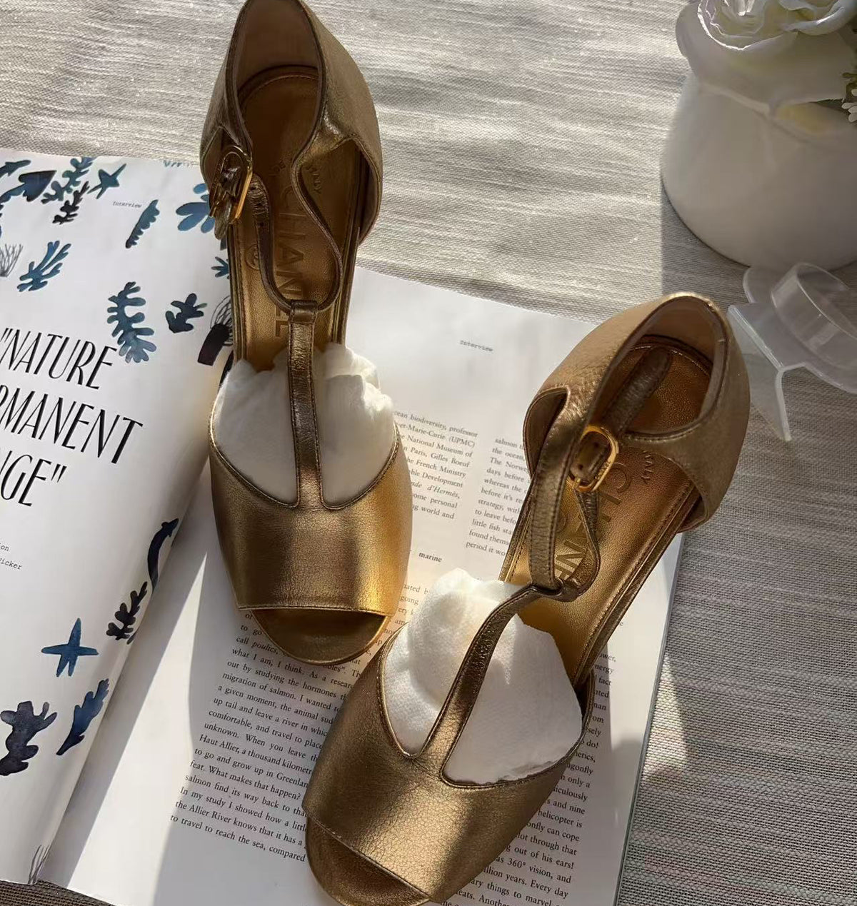 A pair of metallic gold peep-toe high heels with T-strap buckles, displayed on an open book with white tissue paper inserts. A white ceramic vase with flowers is visible in the background on a textured light gray fabric surface.