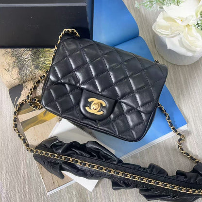 Black quilted Chanel handbag with gold chain on a textured surface with flowers and a box in the background.