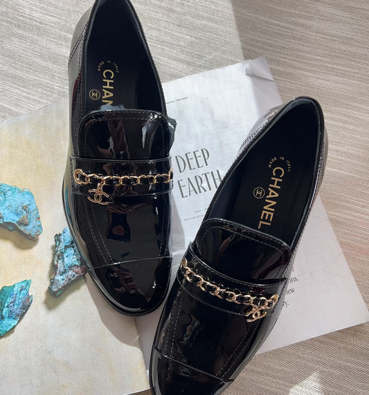 Black patent leather loafers with gold chain detail on a textured surface