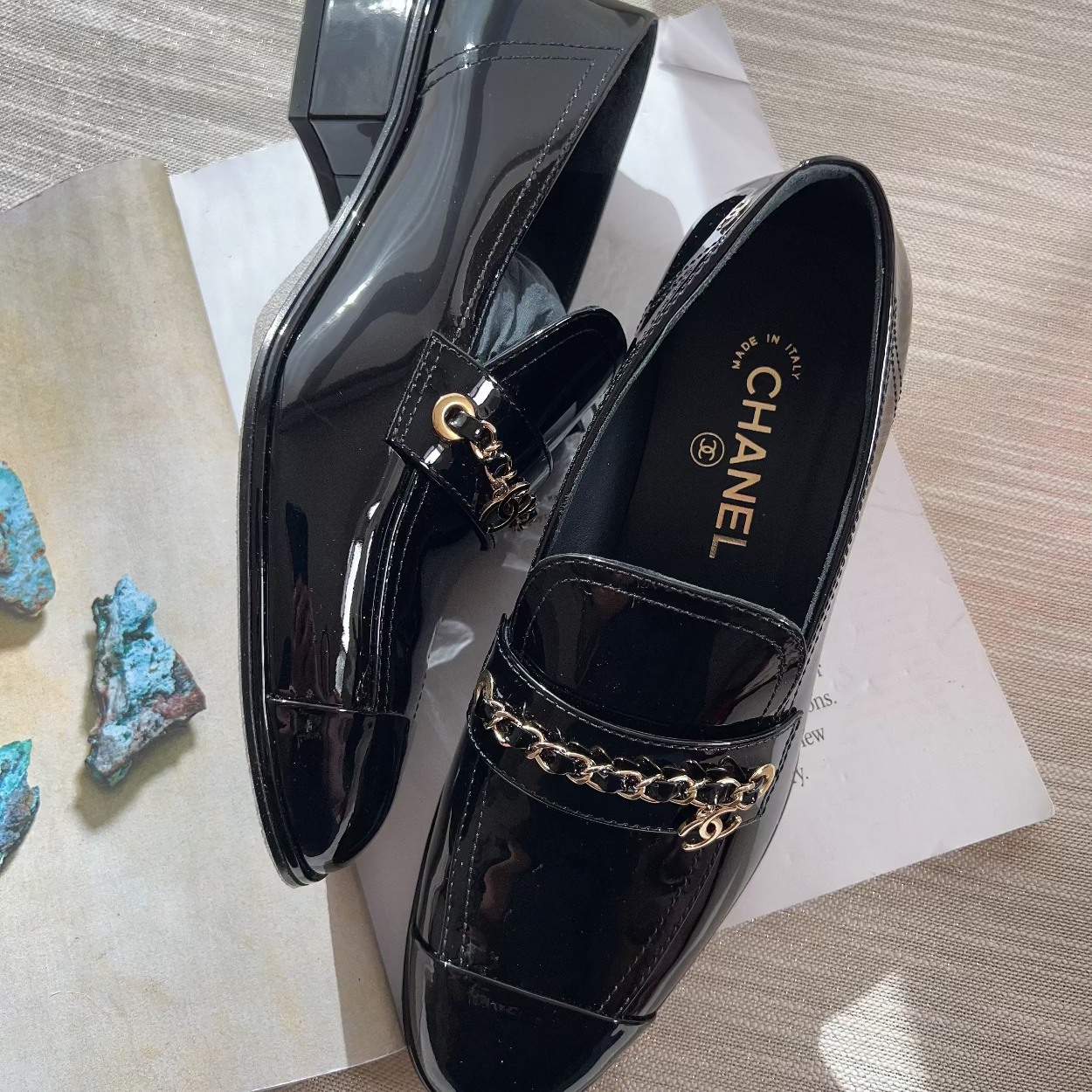 Black patent leather loafers with gold chain detail on a textured surface