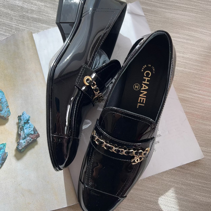 Black patent leather loafers with gold chain detail on a textured surface