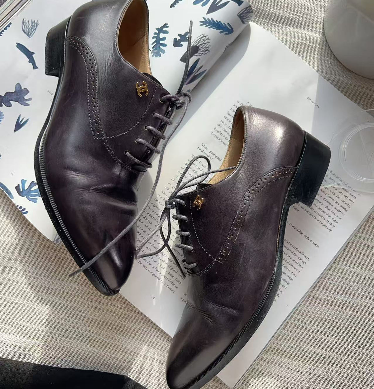 A pair of dark brown leather lace-up oxford shoes with low stacked heels and gold-tone hardware, styled on top of an open magazine over a light wood surface.
