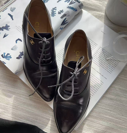 A pair of dark brown leather lace-up oxford shoes with gold-tone logo hardware, displayed on top of an open book.