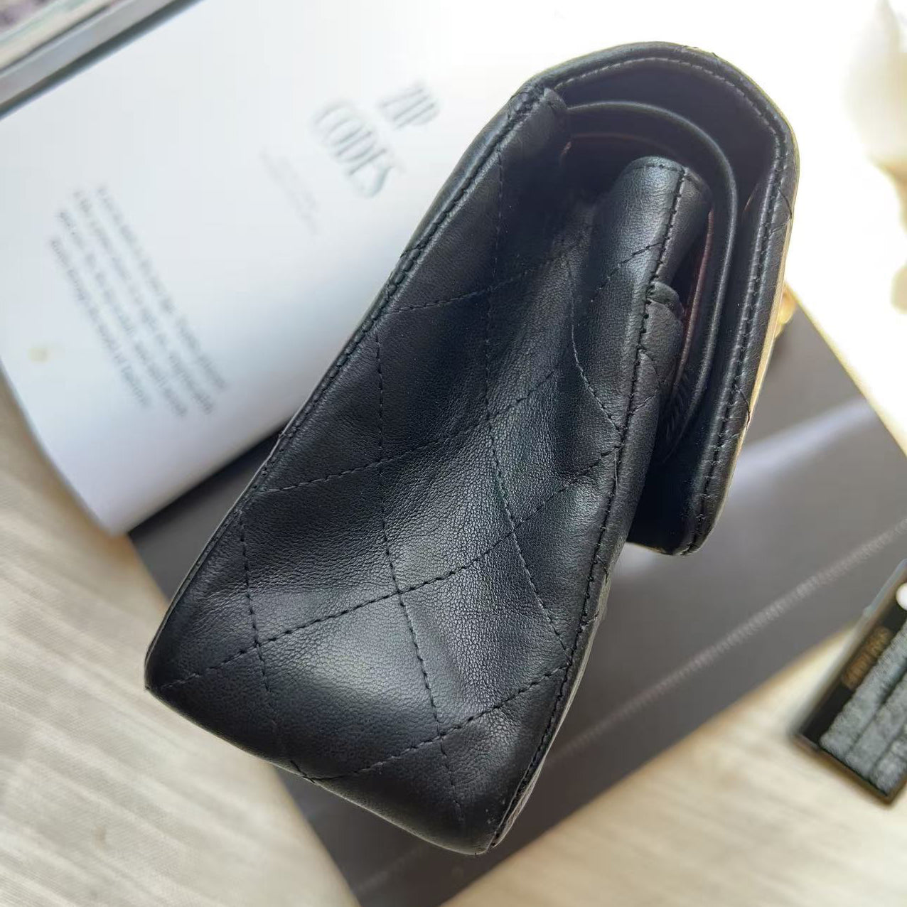 A black quilted leather wallet or card case with a triangular pointed end, photographed on top of a white book and a dark box on a light wood surface.