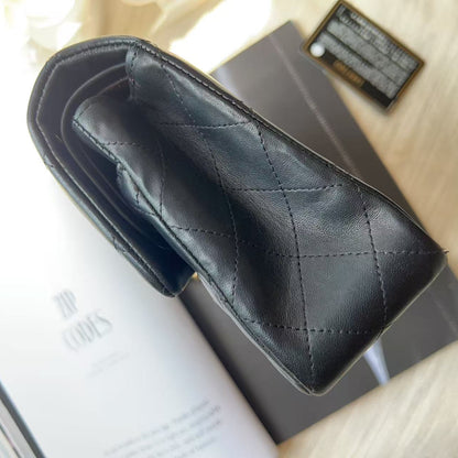 A black quilted leather wallet or card holder shown from an overhead angle, resting on a gray book or box with a magazine underneath on a light beige textured surface.