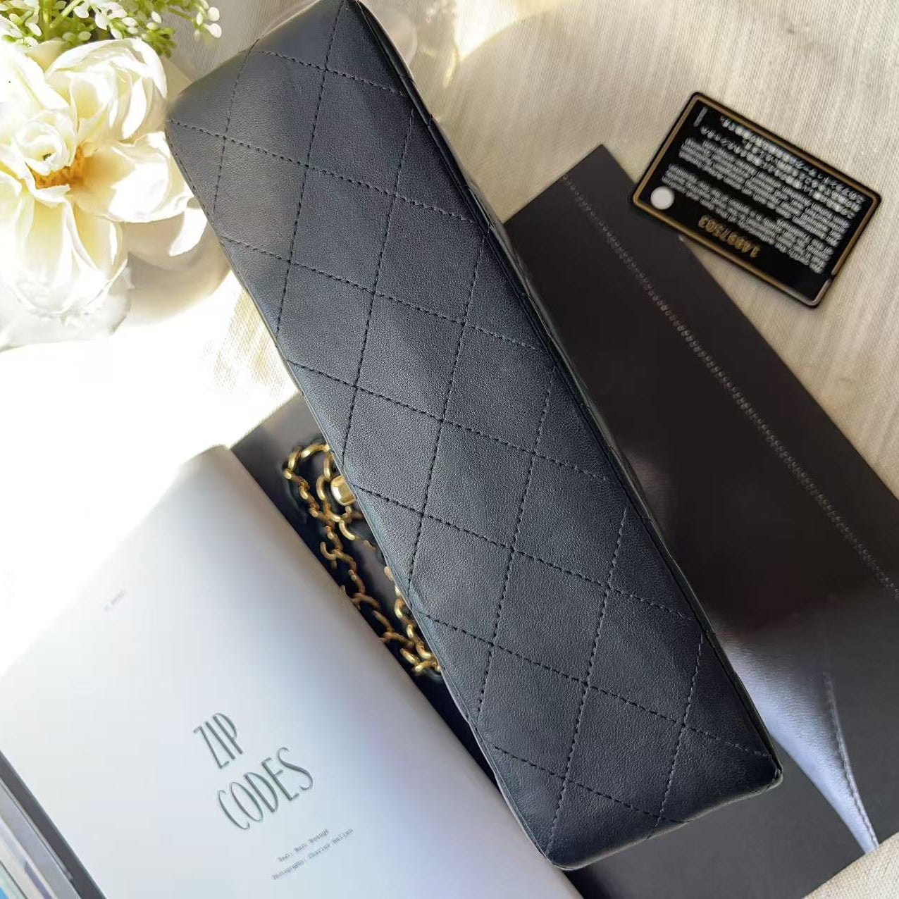 A black quilted leather zip-around wallet or clutch with a gold chain link detail, displayed on top of its black packaging and a white booklet on a light wood surface with white flowers in the corner.