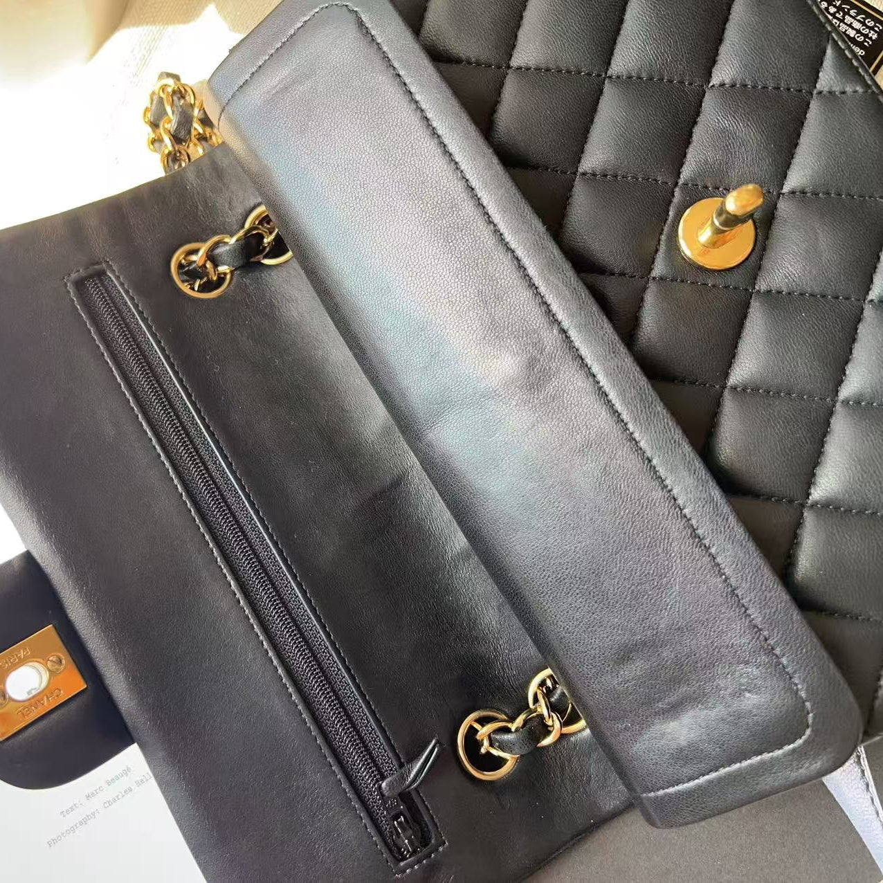 A close-up of a black leather shoulder bag with gold-tone hardware, showing the interior zipper pocket and flap, laid next to a quilted black bag on a light surface.