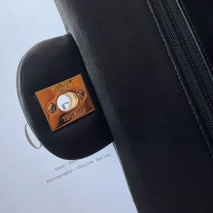 Close-up of a black leather Louis Vuitton bag with a distinctive square bronze-toned metal lock and keyhole detail, set against a light blue background with a printed page.
