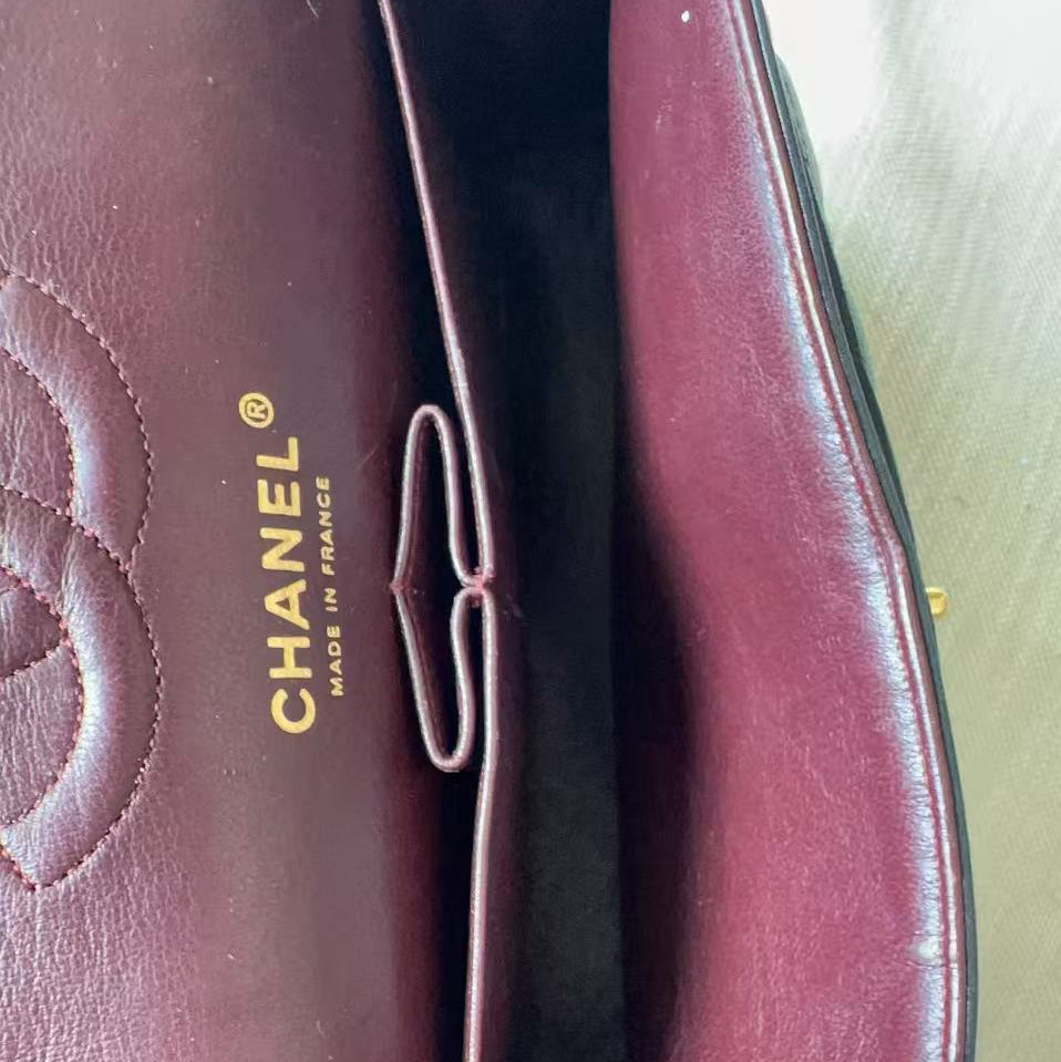 Close-up of a Chanel handbag interior in burgundy leather, showing the gold-embossed CHANEL logo and a finger holding the open flap.