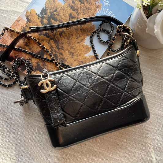 Black quilted handbag with gold chain strap on a wooden surface with a magazine and flowers in the background.