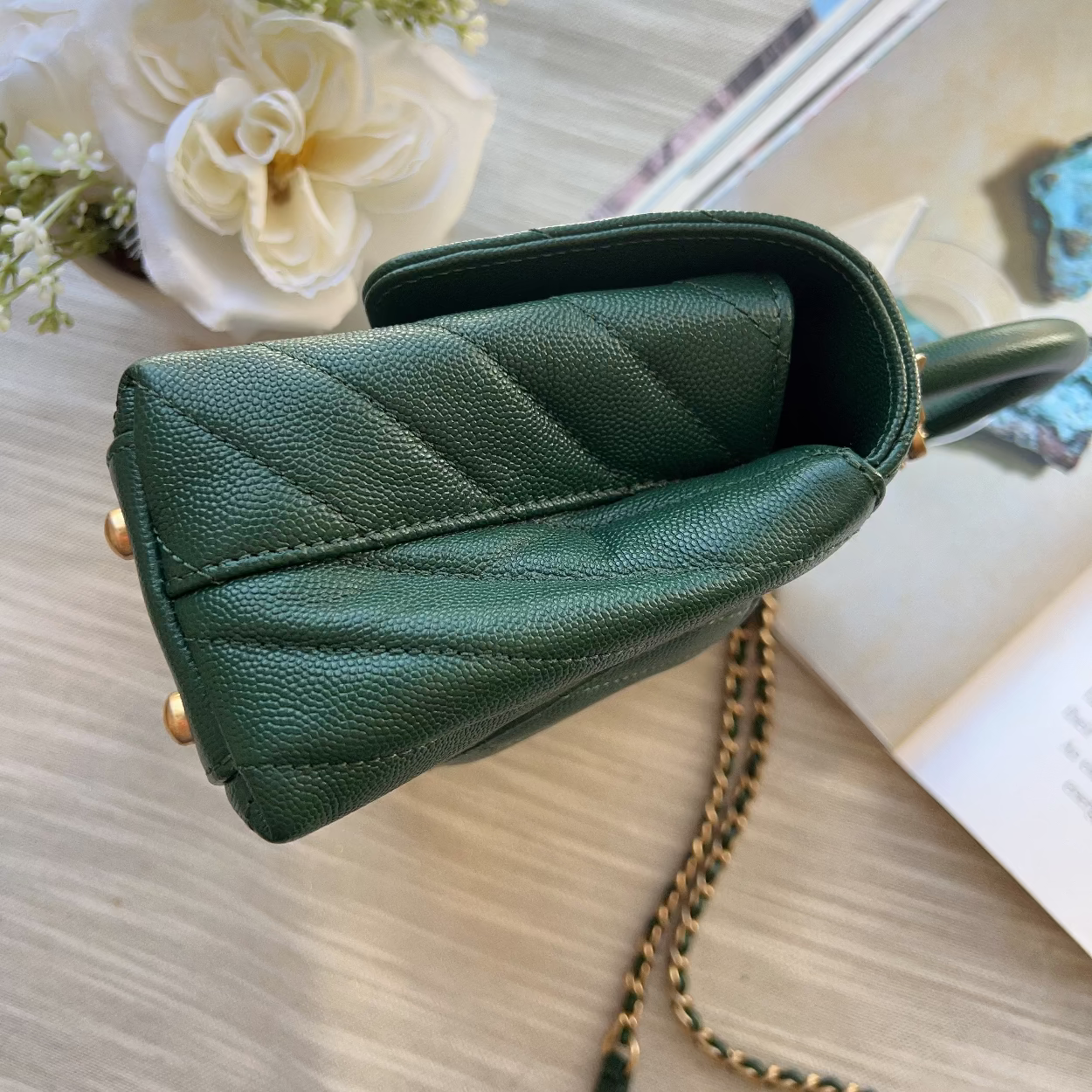 Green handbag with a chain detail on a light surface with flowers and books.