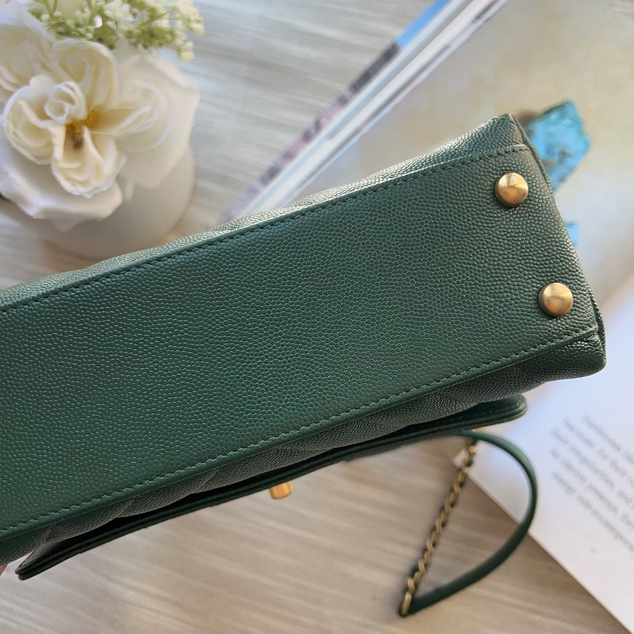 Green wallet with gold studs on a light wooden surface with flowers and a book.