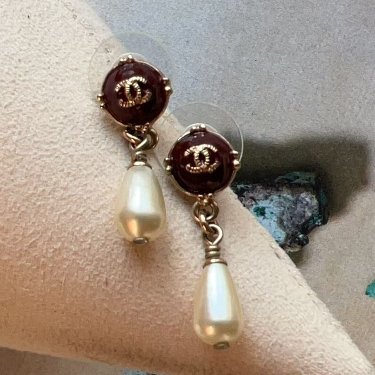 A pair of Chanel drop earrings with dark red enamel studs featuring the interlocking CC logo in gold, hanging from clear posts with cream pearl dangles, displayed on a beige jewelry stand.