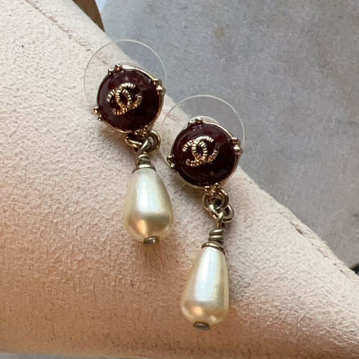 A pair of Chanel drop earrings with dark red circular studs featuring the interlocking CC logo, hanging from gold-toned rings with teardrop-shaped white faux-pearl drops, displayed on a beige jewelry stand.