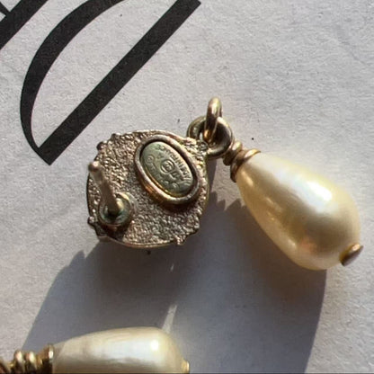 A close-up of a vintage gold-toned earring with a textured oval setting and a dangling cream-colored pearl bead, resting on a white surface with large black letters in the background.