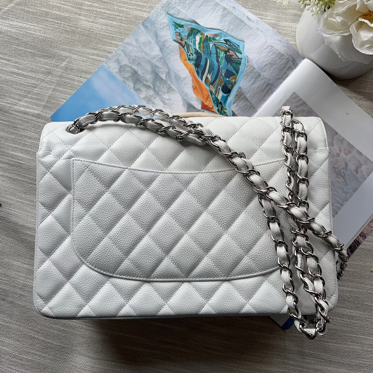 White quilted handbag with a chain strap on a textured surface with a magazine and flowers.