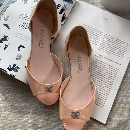 A pair of Chanel peep-toe slingback flats in a glossy nude pink patent leather, featuring the iconic interlocking CC logo on the toe. The shoes are displayed on a gray textured fabric surface, styled with open fashion magazines.
