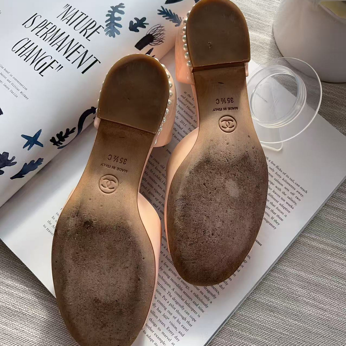 A pair of women's low-heeled sandals shown from the sole side, resting on an open magazine over a light wood surface. The soles are tan with a circular logo stamp and a row of small clear rhinestones along the edge.