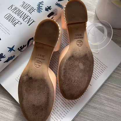 A pair of women's low-heeled sandals shown from the sole side, resting on an open magazine over a light wood surface. The soles are tan with a circular logo stamp and a row of small clear rhinestones along the edge.