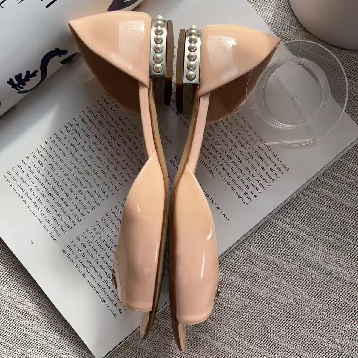 A pair of glossy nude beige kitten heel pumps with silver rhinestone-embellished straps, arranged on an open book with a glass and a decorative bag nearby.