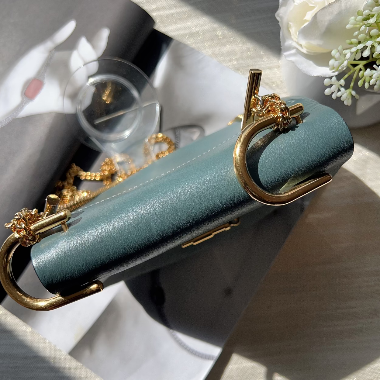 Teal handbag with gold clasp on a wooden surface with flowers in the background