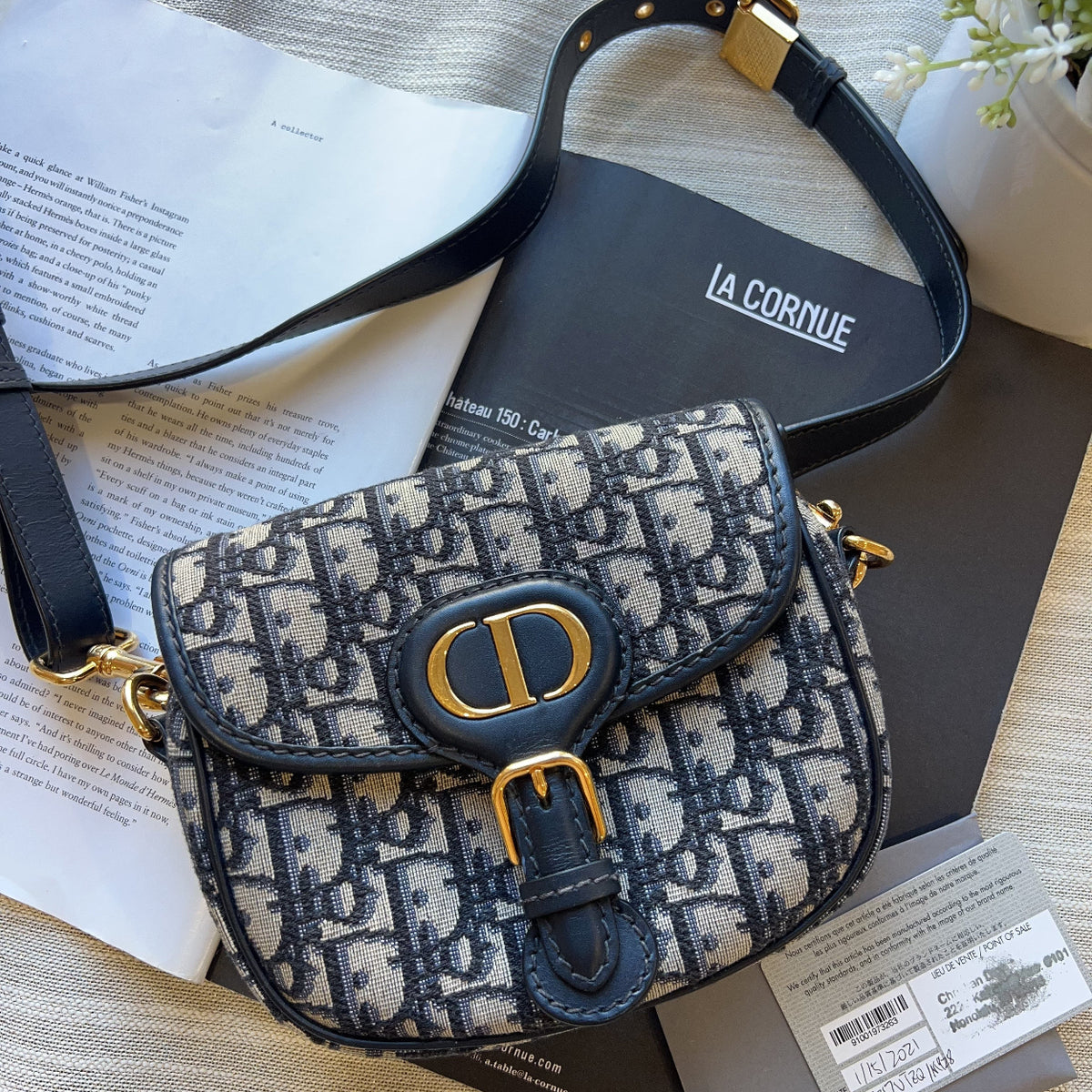 A Dior Saint Laurent shoulder bag in the iconic Oblique monogram canvas, featuring navy blue leather trim and gold-tone hardware, displayed on a textured beige fabric with its dust bag and papers.