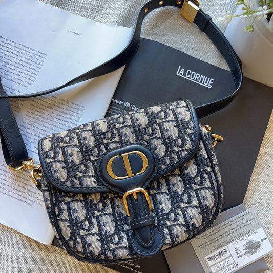 A Dior Saint Laurent shoulder bag in the iconic Oblique monogram canvas, featuring navy blue leather trim and gold-tone hardware, displayed on a textured beige fabric with its dust bag and papers.