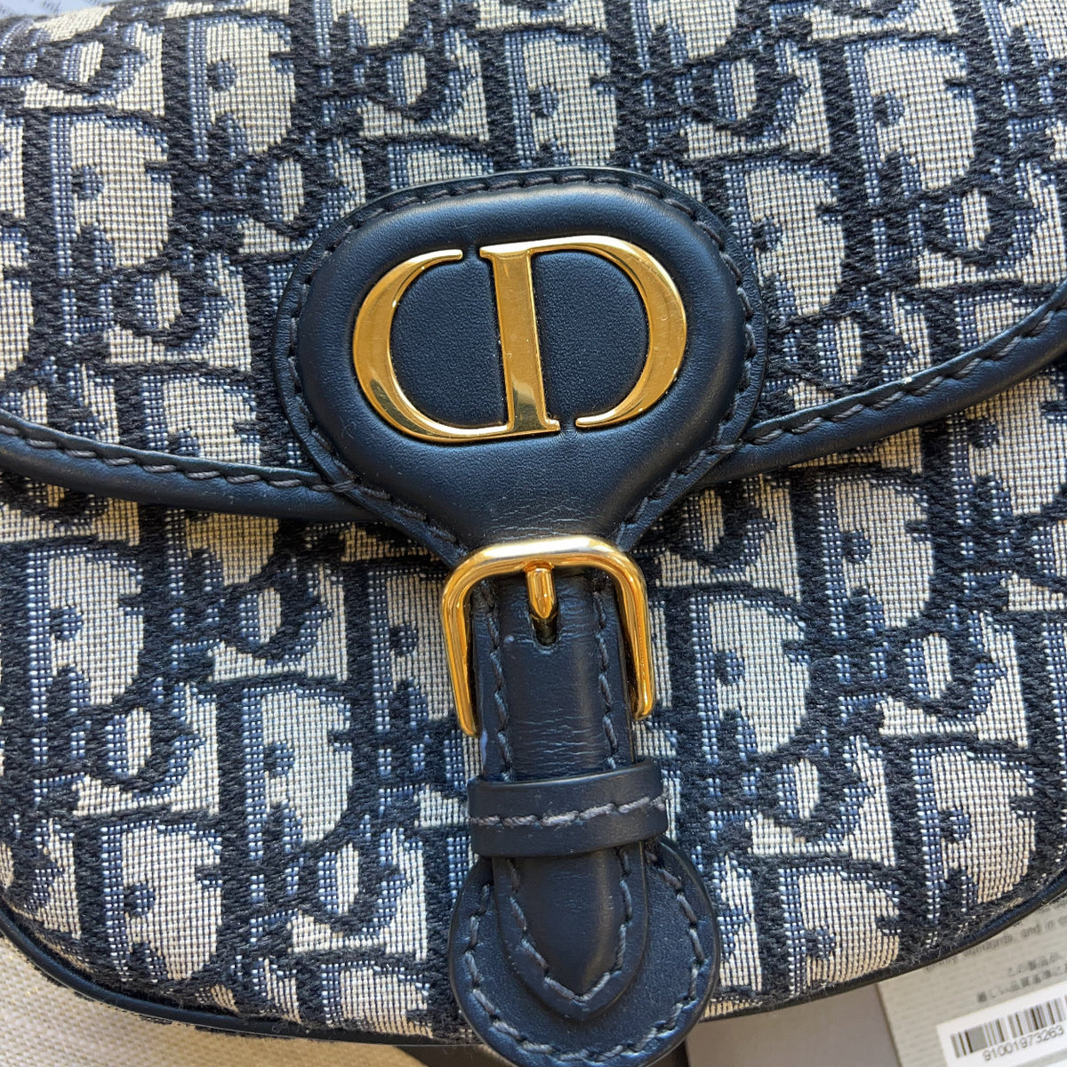 Close-up of a Dior monogram handbag in navy blue and beige canvas, featuring a navy leather strap with a gold-tone D logo clasp and buckle.