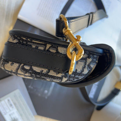 A close-up of a designer handbag strap featuring a black and beige monogram pattern, a glossy black leather section, and a prominent gold-tone metal clasp, resting on a light-colored surface with its packaging box visible in the background.