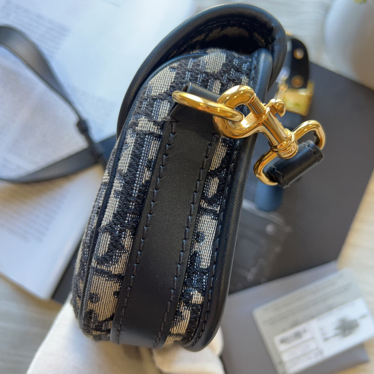 A close-up of a Dior monogram shoulder bag with black leather trim and a gold horsebit clasp, held by a hand wearing a white glove with its packaging and documents in the background.