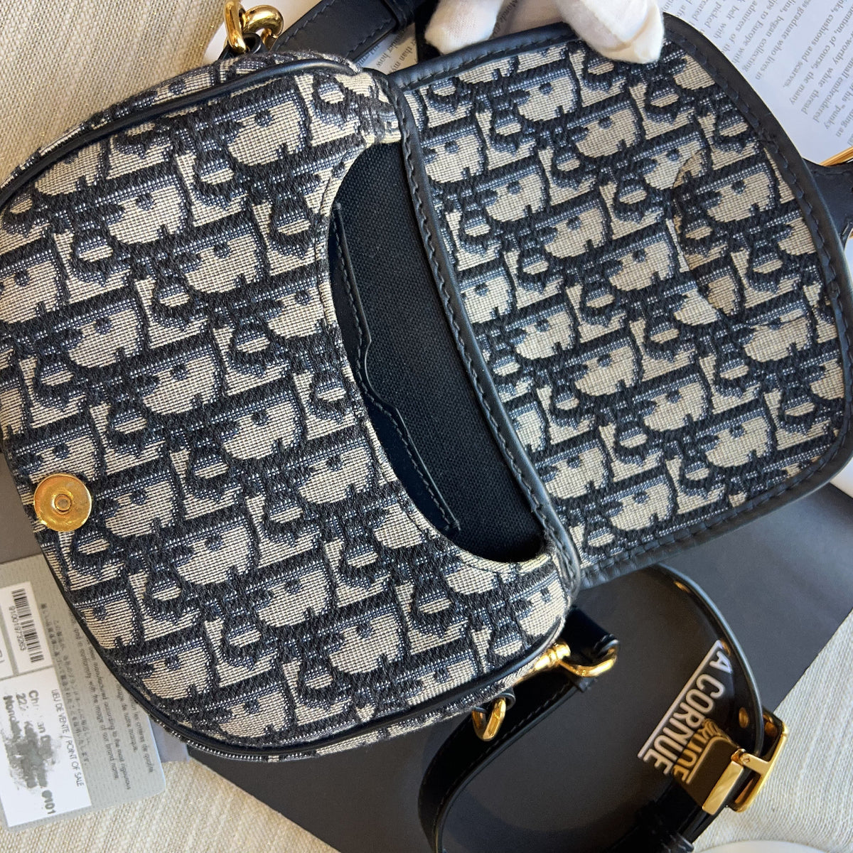 A Dior Oblique canvas shoulder bag with black leather trim and gold-tone hardware, shown with its original tag and a black Dior strap on a light wood surface.