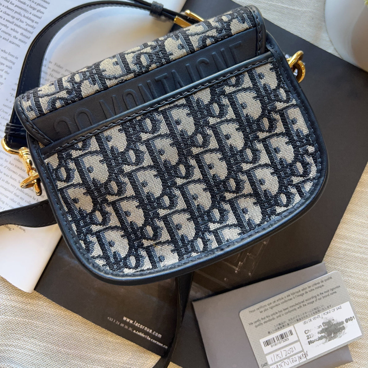 A Dior Saddle bag in navy blue and beige monogram canvas, displayed on a dark surface with its original box and papers.