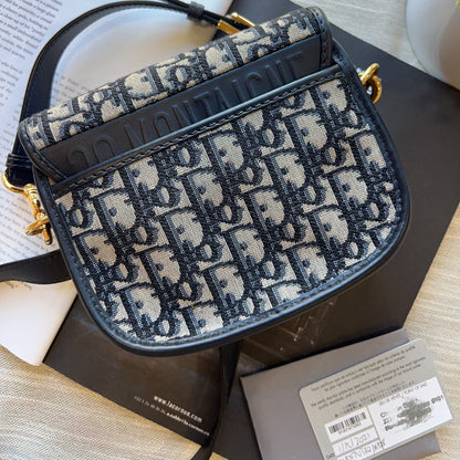 A Dior Saddle bag in navy blue and beige monogram canvas, displayed on a dark surface with its original box and papers.