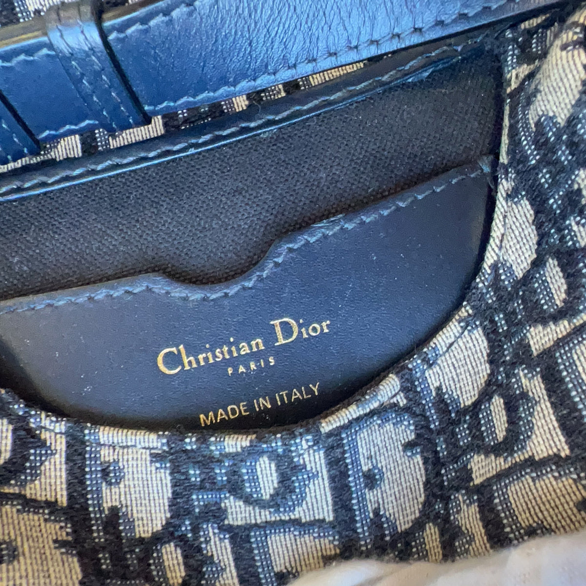 Close-up of a Christian Dior handbag showing the interior lining with gold 'Christian Dior' branding, a blue leather strap, and the signature beige-and-navy monogram canvas. A white-gloved finger is visible touching the bag.