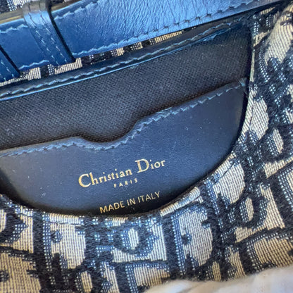 Close-up of a Christian Dior handbag showing the interior lining with gold 'Christian Dior' branding, a blue leather strap, and the signature beige-and-navy monogram canvas. A white-gloved finger is visible touching the bag.
