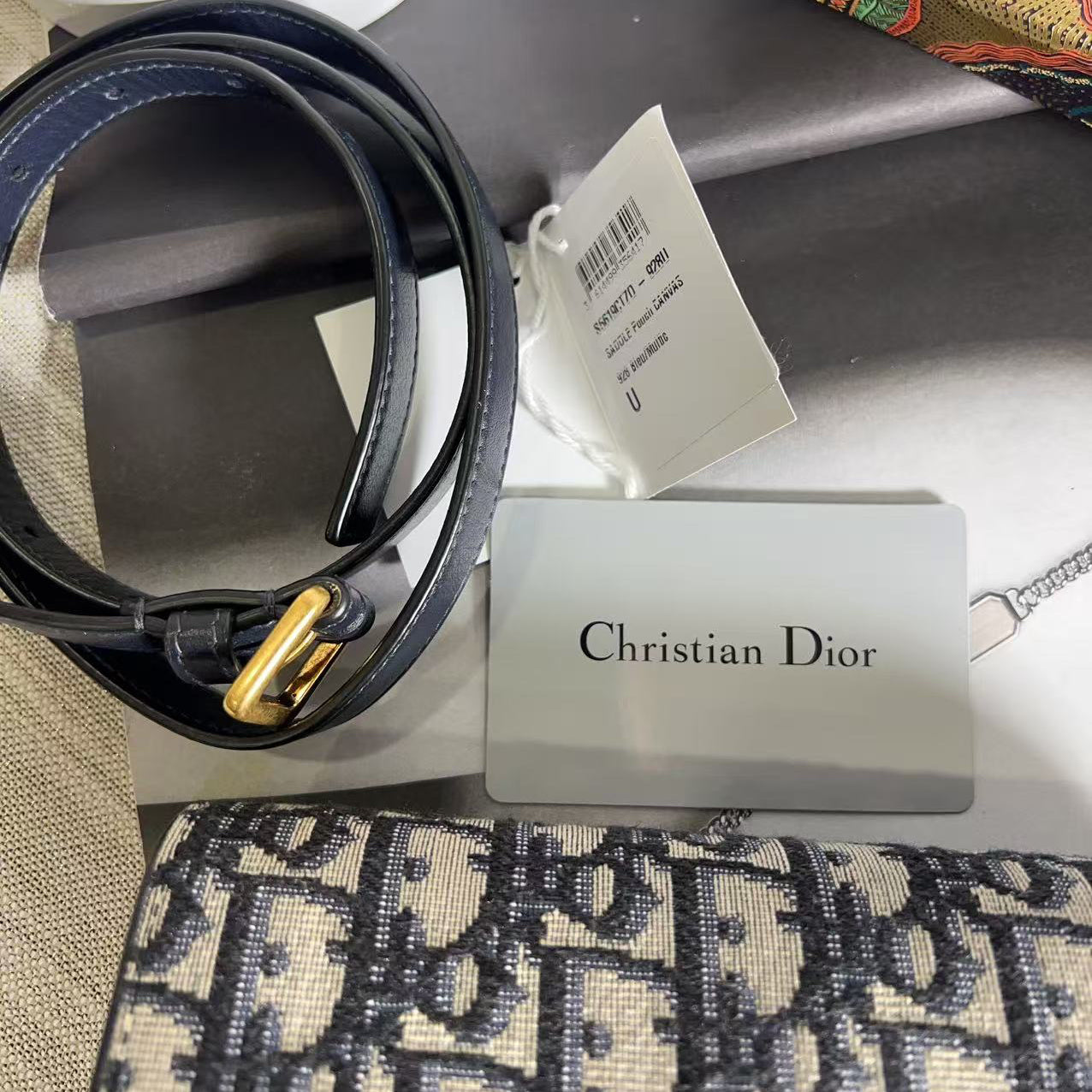 Black belt with gold buckle on a stand with a Christian Dior tag, placed on a patterned fabric surface.