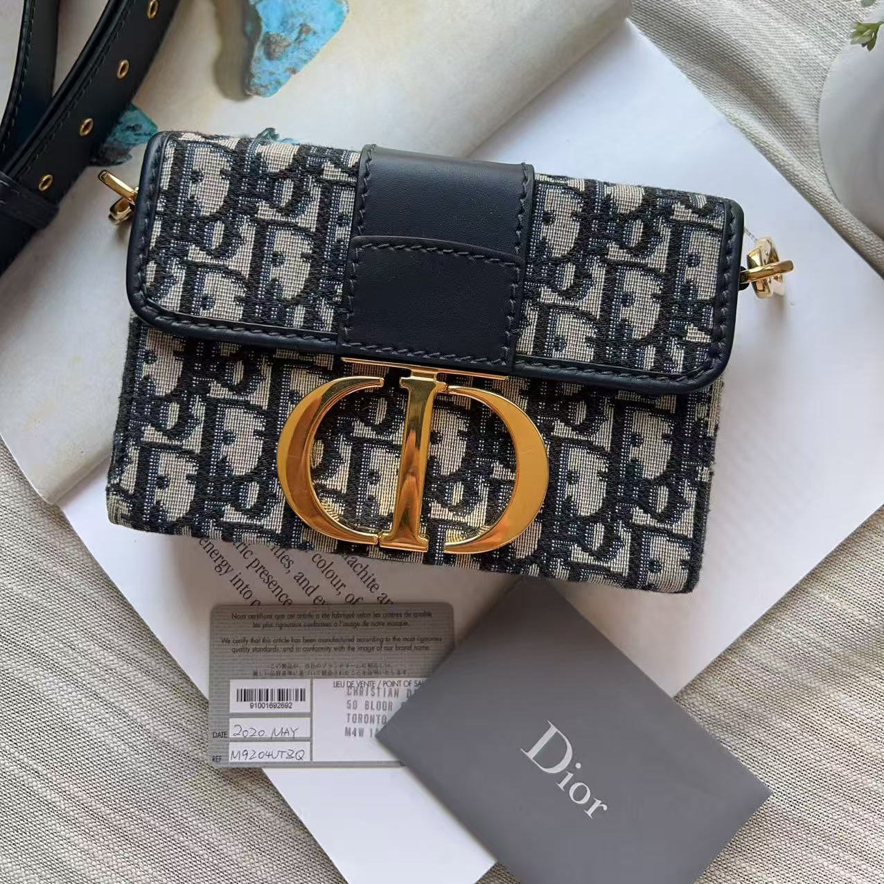 Dior Oblique monogram shoulder bag with a black leather strap and gold-tone hardware, displayed on a white surface with a Dior card and accessories.