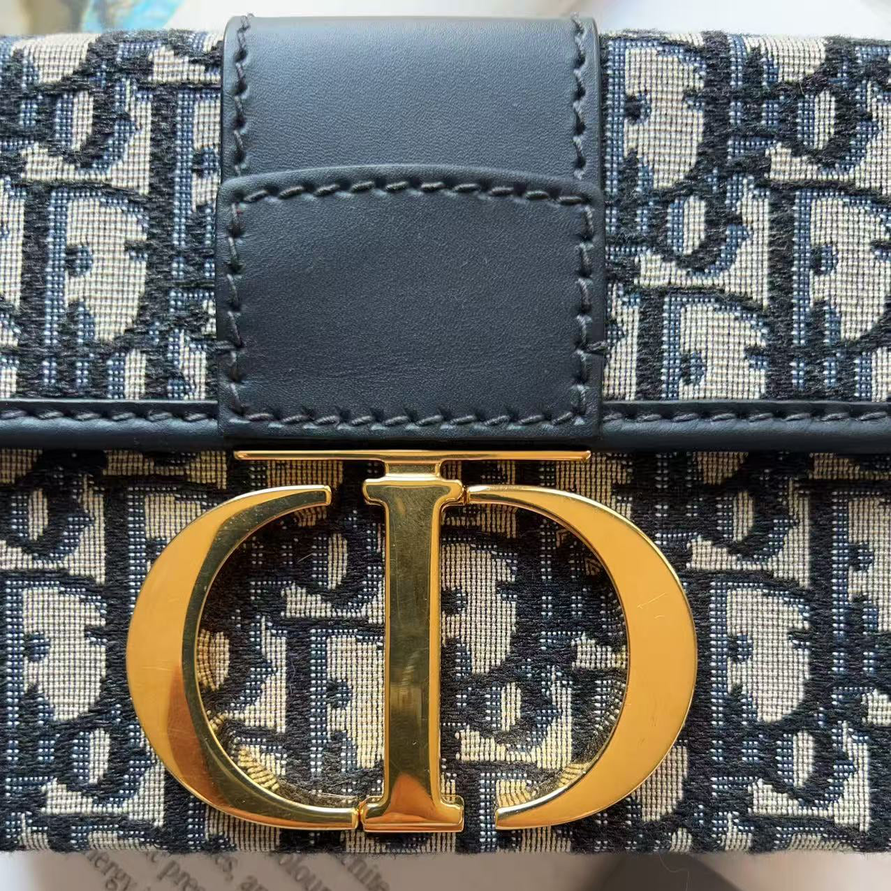 Close-up of a Dior monogram wallet or clutch with a dark navy leather tab and a large gold-tone CD clasp, shown with shopping tags in the background.