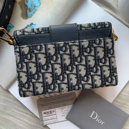 A Dior monogram crossbody bag in navy and beige coated canvas with a dark navy leather top handle and gold-tone hardware, displayed on a light surface with a Dior card and tags nearby.