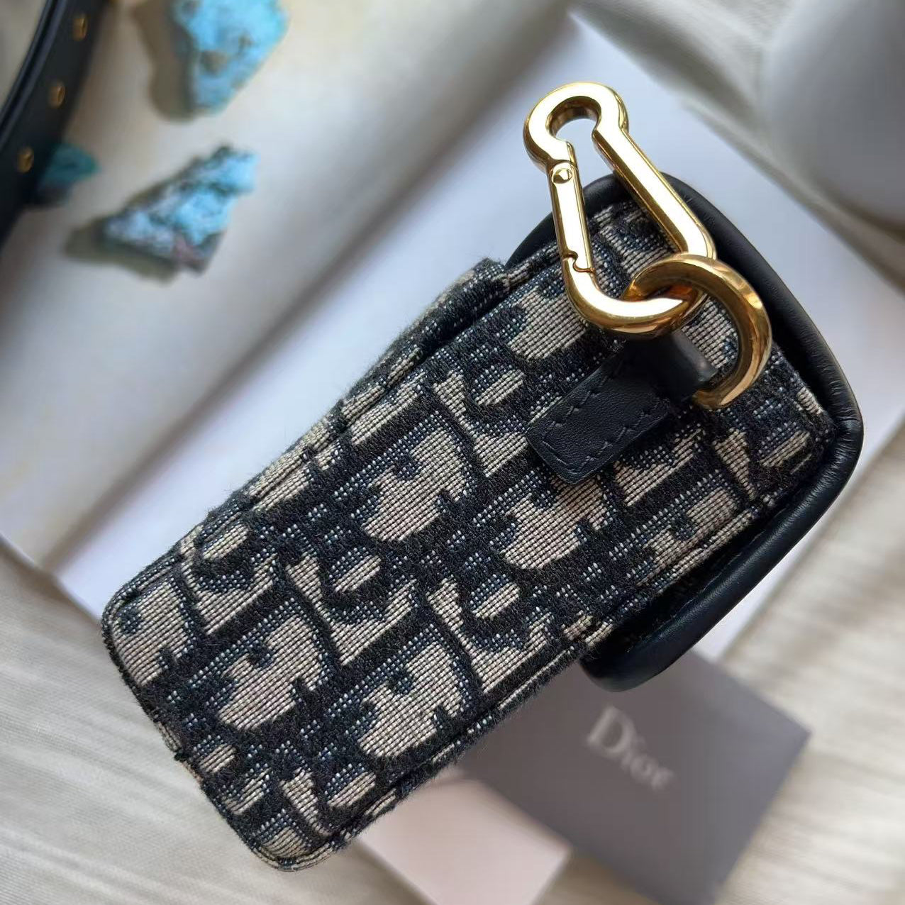 A Dior monogram canvas key fob with a gold-tone carabiner clip, displayed on a light wood surface next to a Dior box.