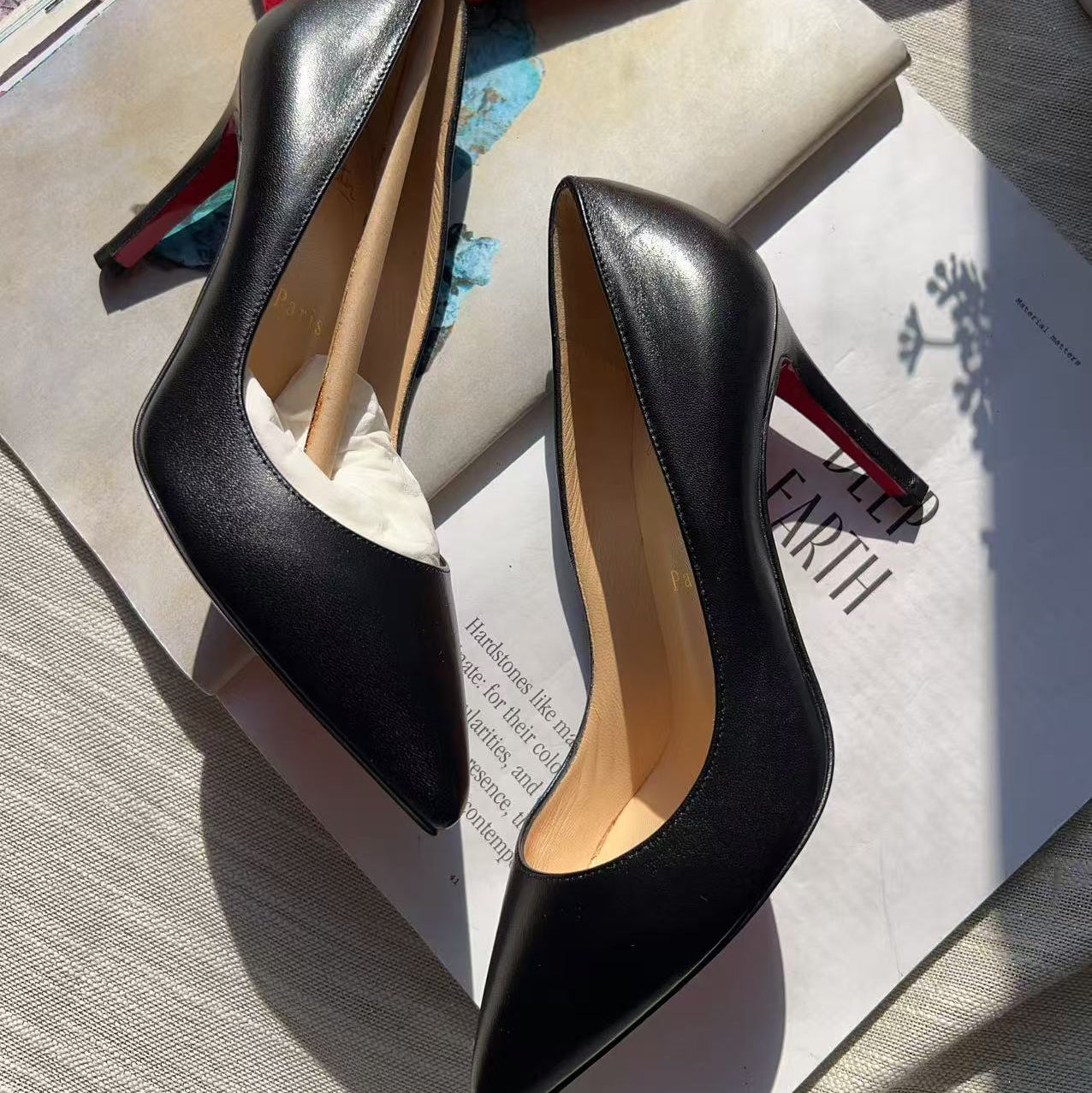 A pair of black leather pointed-toe pump heels with red stiletto heels, displayed on top of a magazine and a red Louis Vuitton dust bag.