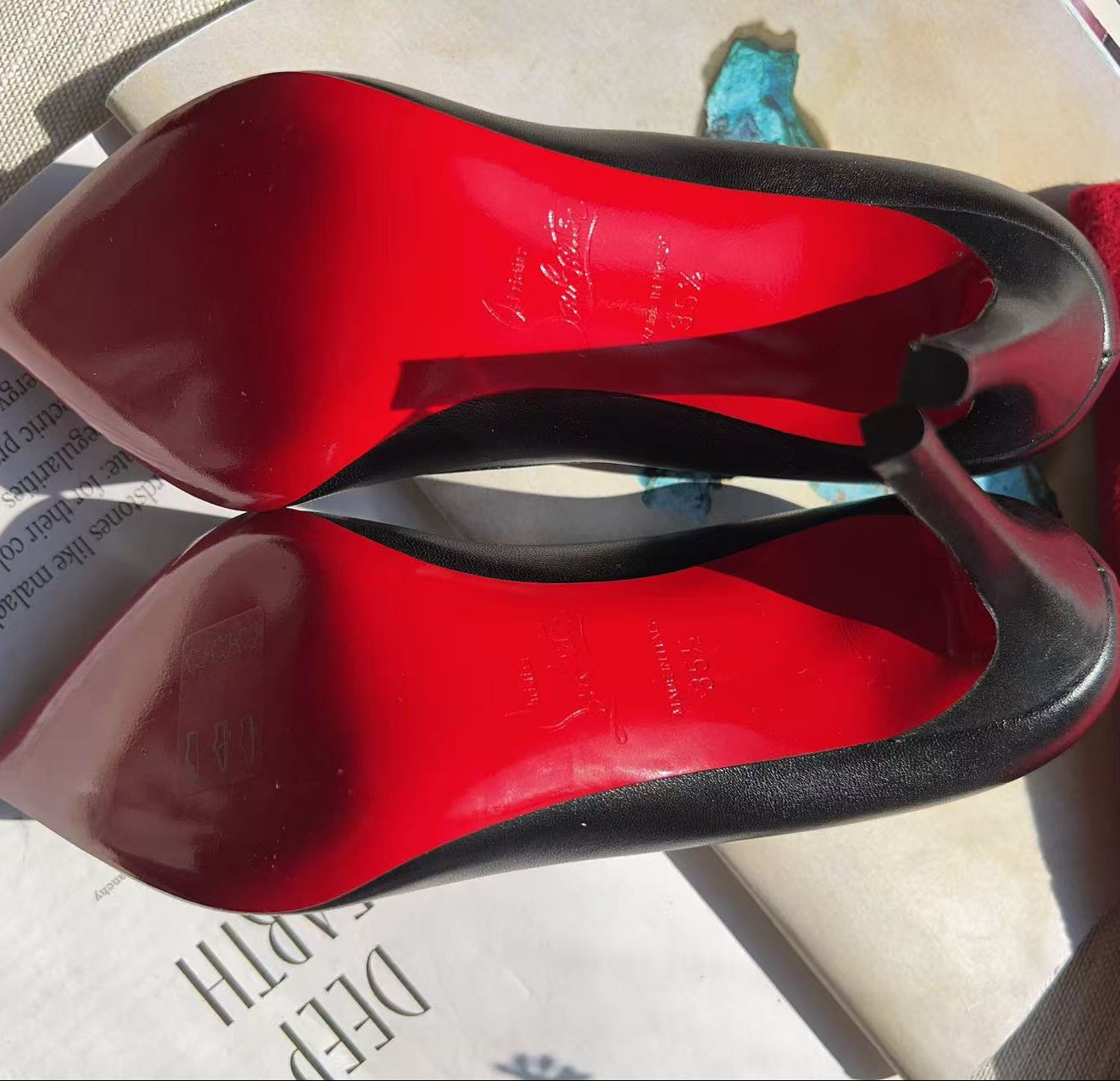 A pair of black pointed-toe high heels with a glossy bright red sole, arranged on top of a book next to a red textured fabric.