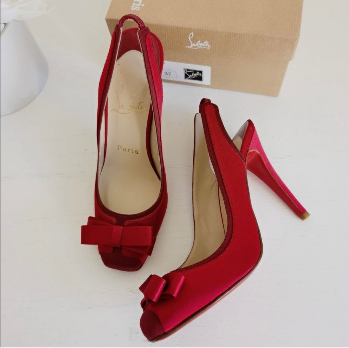 A pair of red satin Christian Louboutin slingback high heels with bow details on the toes, displayed next to their signature brown box on a white surface.